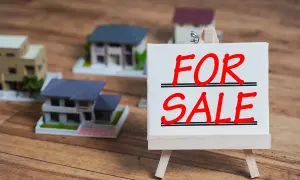Modele domów na drewnianym tle. Tabliczka "FOR SALE" sugeruje, jak sprzedać mieszkanie z hipoteką.