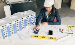 Architektka w kasku analizuje projekt budynku, sprawdzając zgodność z warunkami technicznymi. Na stole makieta, plany, kalkulator i poziomica.