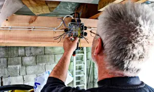 Mężczyzna montuje prostą instalację elektryczną w garażu. Widać skrzynkę z przewodami i narzędzia.