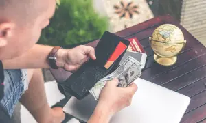 Mężczyzna liczy pieniądze w portfelu, planując sprzedaż mieszkania z kredytem i kupno nowego. Na stole leży globus i laptop.