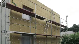 Budynek w trakcie termomodernizacji. Elewacja ocieplana wełną mineralną, otoczona rusztowaniem. Widoczne okna i drabiny.
