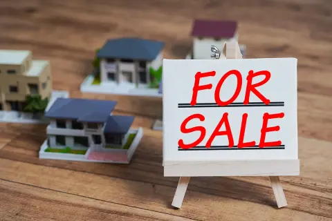 Modele domów na drewnianym tle. Tabliczka "FOR SALE" sugeruje, jak sprzedać mieszkanie z hipoteką.