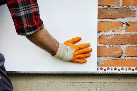 Pracownik w pomarańczowych rękawicach przykłada styropian do ceglanej ściany. Zastanawiasz się, ile kosztuje ankrowanie domu?