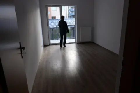Mężczyzna w pustym mieszkaniu, zastanawia się, gdzie złożyć wniosek o mieszkanie komunalne. Widok na balkon i sąsiednie budynki.