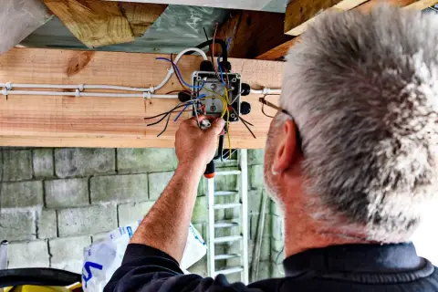 Mężczyzna montuje prostą instalację elektryczną w garażu. Widać skrzynkę z przewodami i narzędzia.