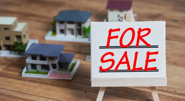 Modele domów na drewnianym tle. Tabliczka "FOR SALE" sugeruje, jak sprzedać mieszkanie z hipoteką.
