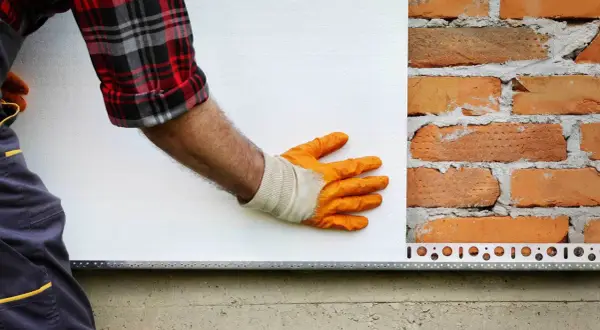 Pracownik w pomarańczowych rękawicach przykłada styropian do ceglanej ściany. Zastanawiasz się, ile kosztuje ankrowanie domu?
