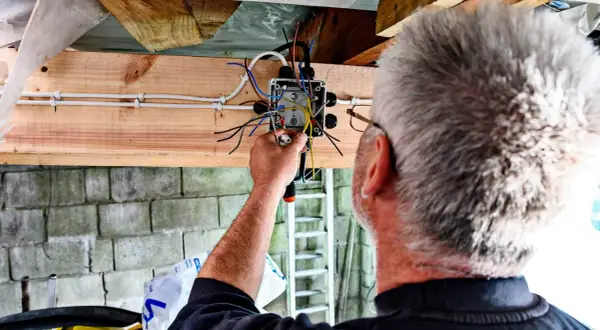 Mężczyzna montuje prostą instalację elektryczną w garażu. Widać skrzynkę z przewodami i narzędzia.