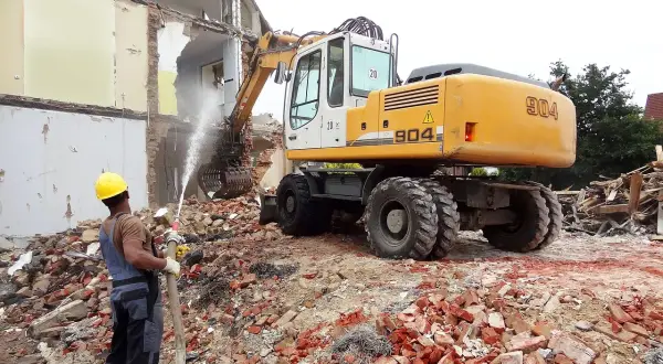 Robot budowlany i pracownik z wężem wodnym na gruzowisku. Zastanawiasz się, ile kosztuje wyburzenie domu?