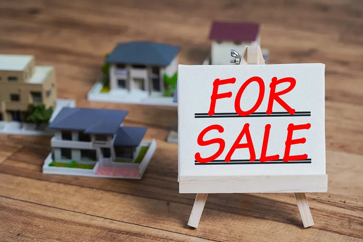 Modele domów na drewnianym tle. Tabliczka "FOR SALE" sugeruje, jak sprzedać mieszkanie z hipoteką.