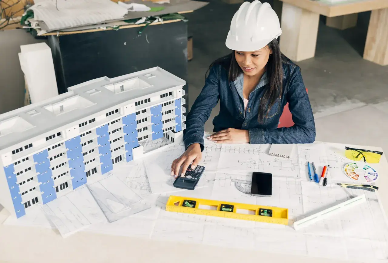 Architektka w kasku analizuje projekt budynku, sprawdzając zgodność z warunkami technicznymi. Na stole makieta, plany, kalkulator i poziomica.