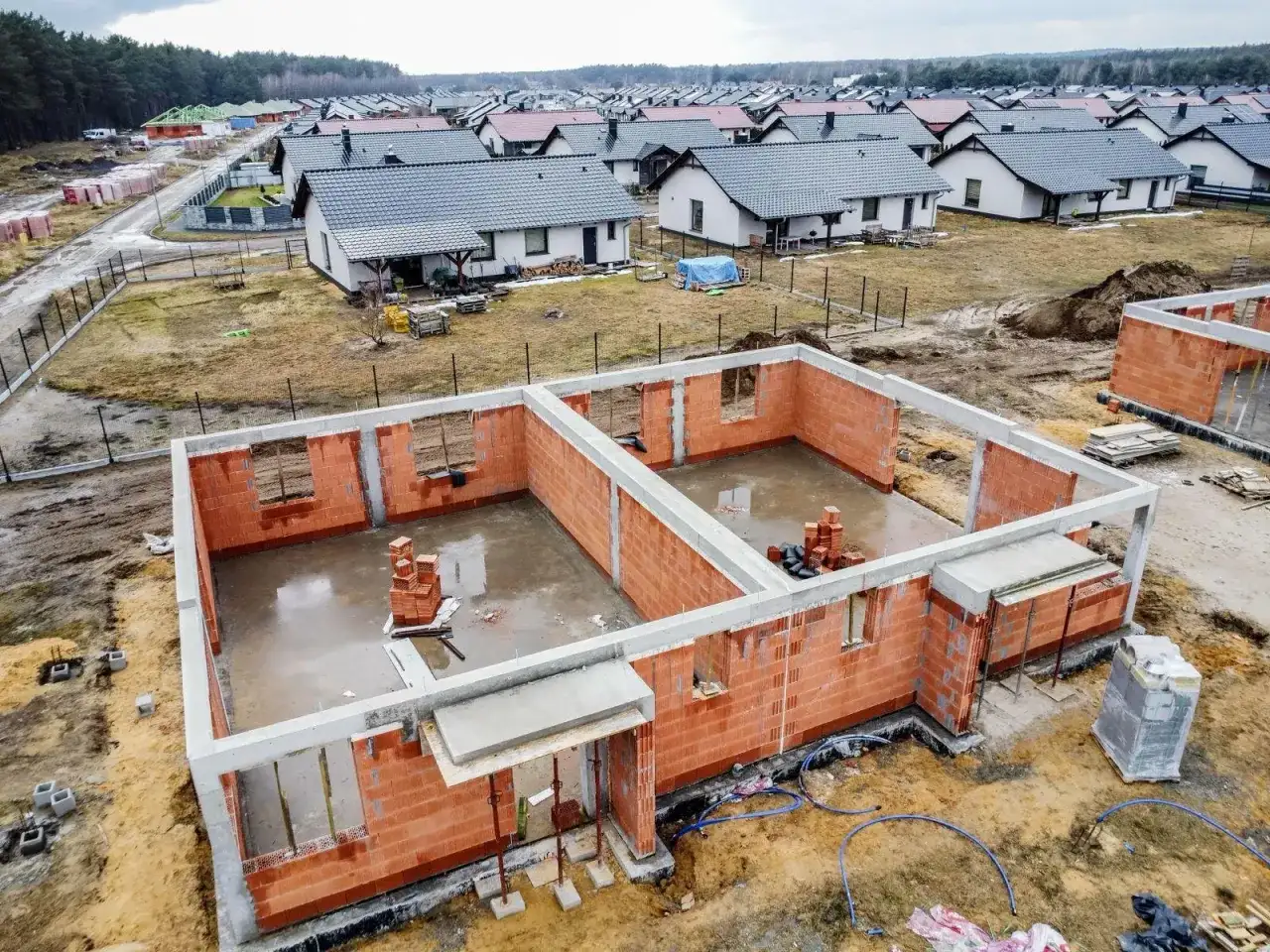 Budowa domu od podstaw, jak zostać deweloperem. Fundamenty i ściany z cegły, w tle osiedle gotowych domów.