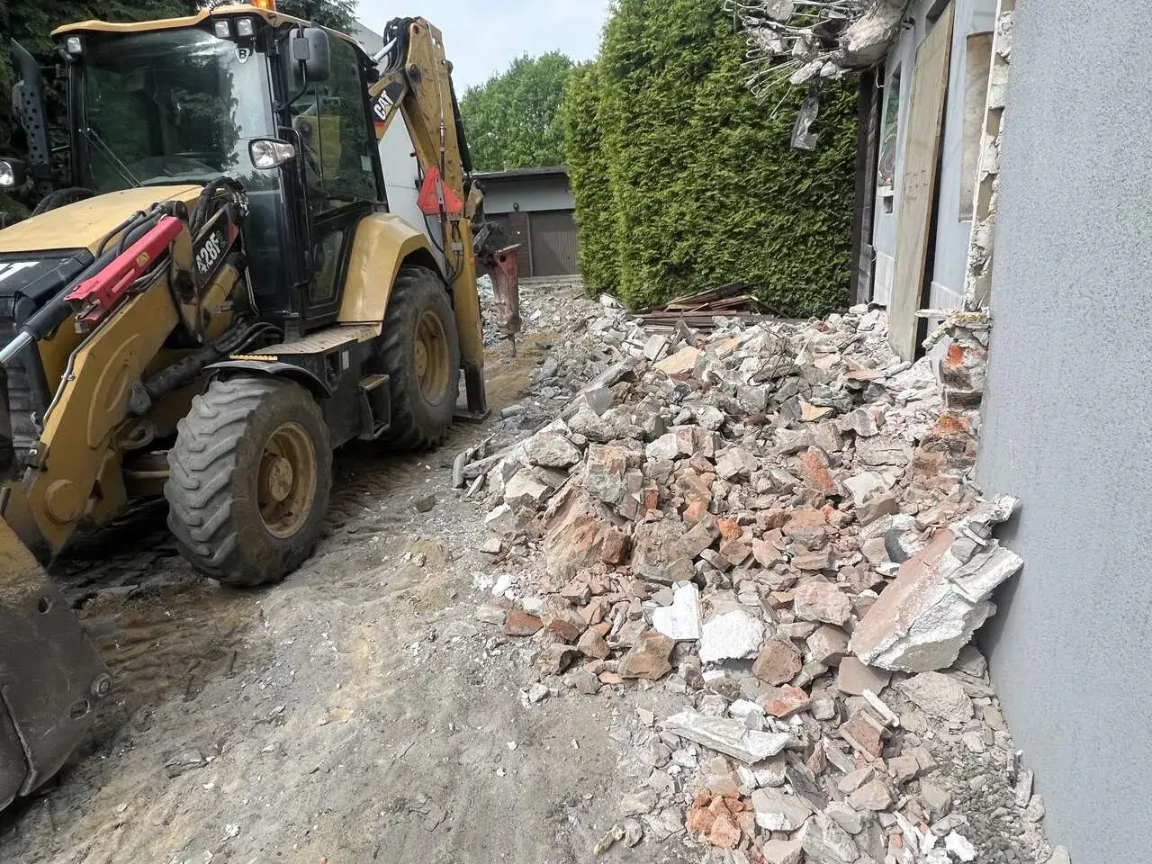 Żółta koparko-ładowarka pracuje przy rozbiórce domu. Gruz i cegły leżą na ziemi. Zastanawiasz się, ile kosztuje wyburzenie domu?