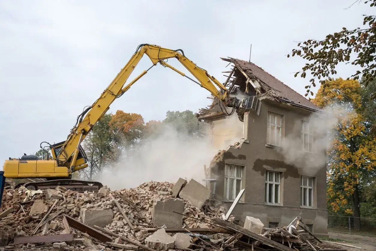 Żółta koparka burzy dom, tworząc chmurę pyłu. Zastanawiasz się, ile kosztuje wyburzenie domu?