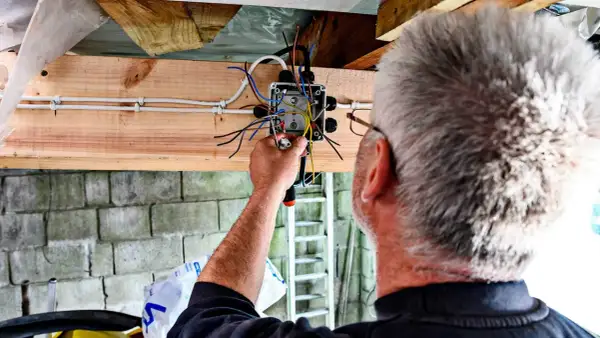 Mężczyzna montuje prostą instalację elektryczną w garażu. Widać skrzynkę z przewodami i narzędzia.